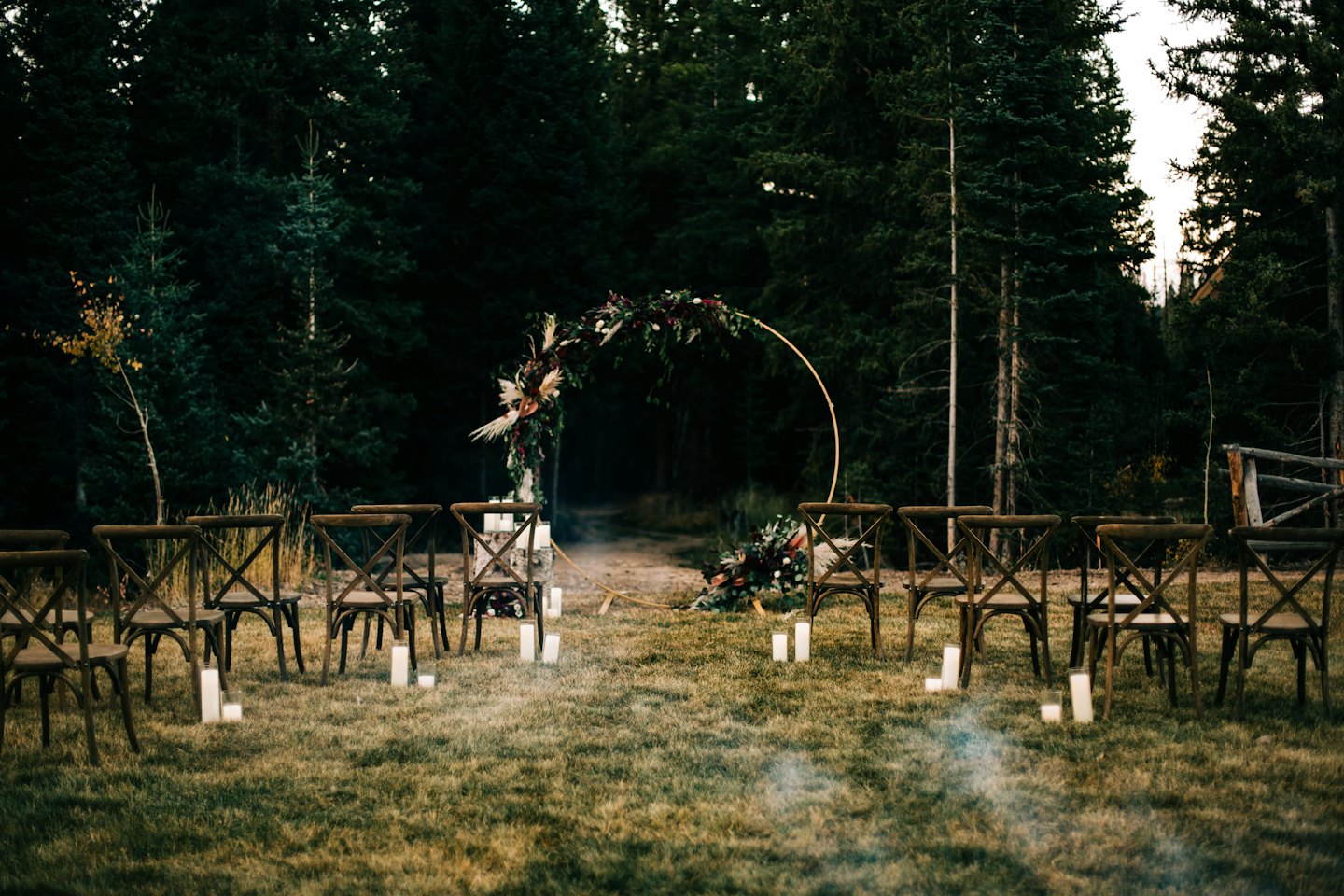 Outdoor wedding ceremony setup with chairs, floral arch, and candles in a forest.